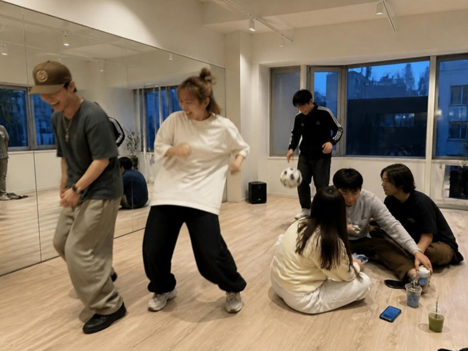 白い壁と明るい木の床のスタジオで、笑いながら踊る二人、床に座って話す人たち、奥でボールを扱う人（イメージ）
